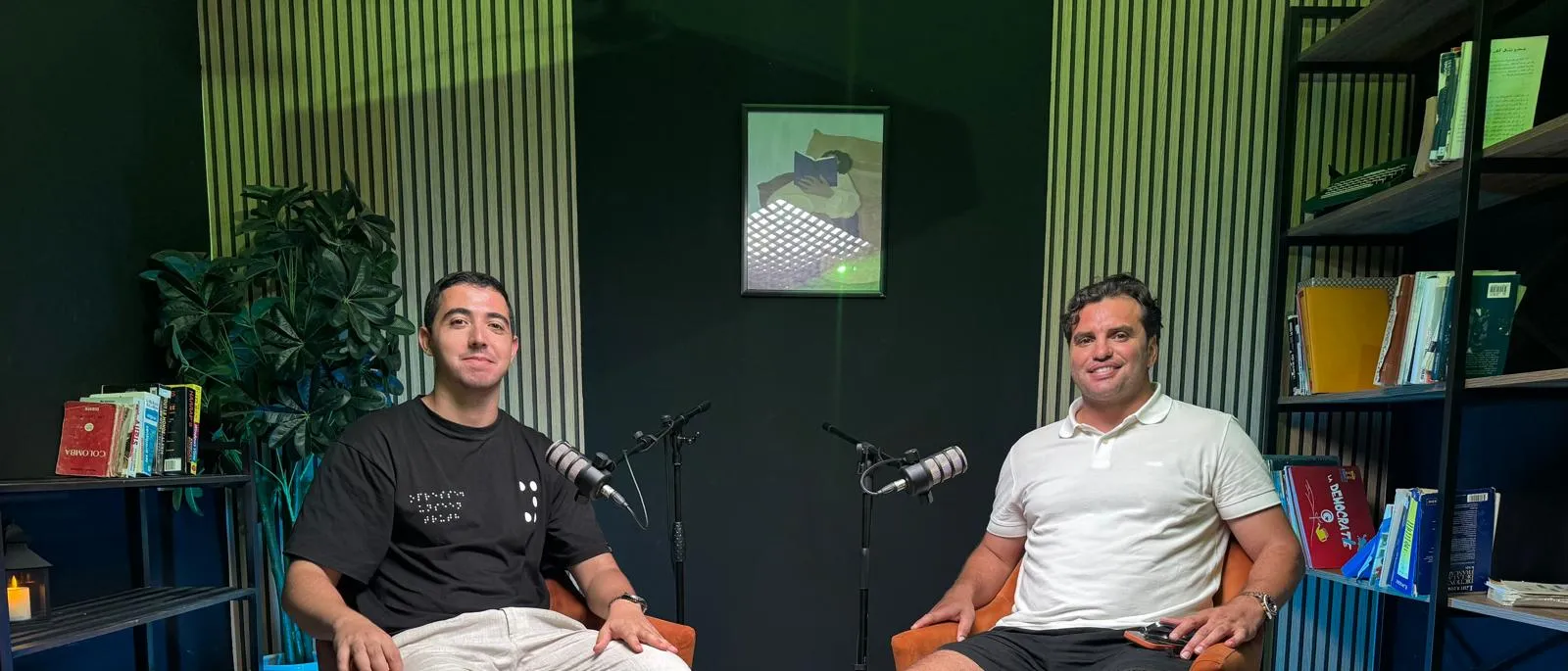 Two men seated in orange armchairs at podcast microphones in a dark, slatted-wood studio lined with books
